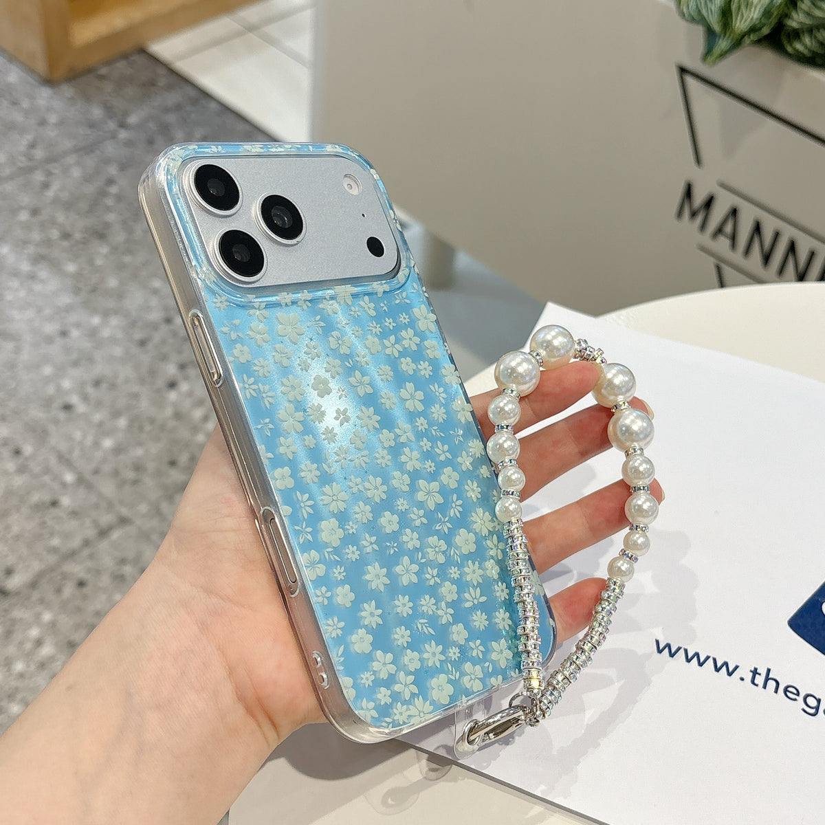 Blue floral phone case with a pearl chain held by a hand on a white surface.