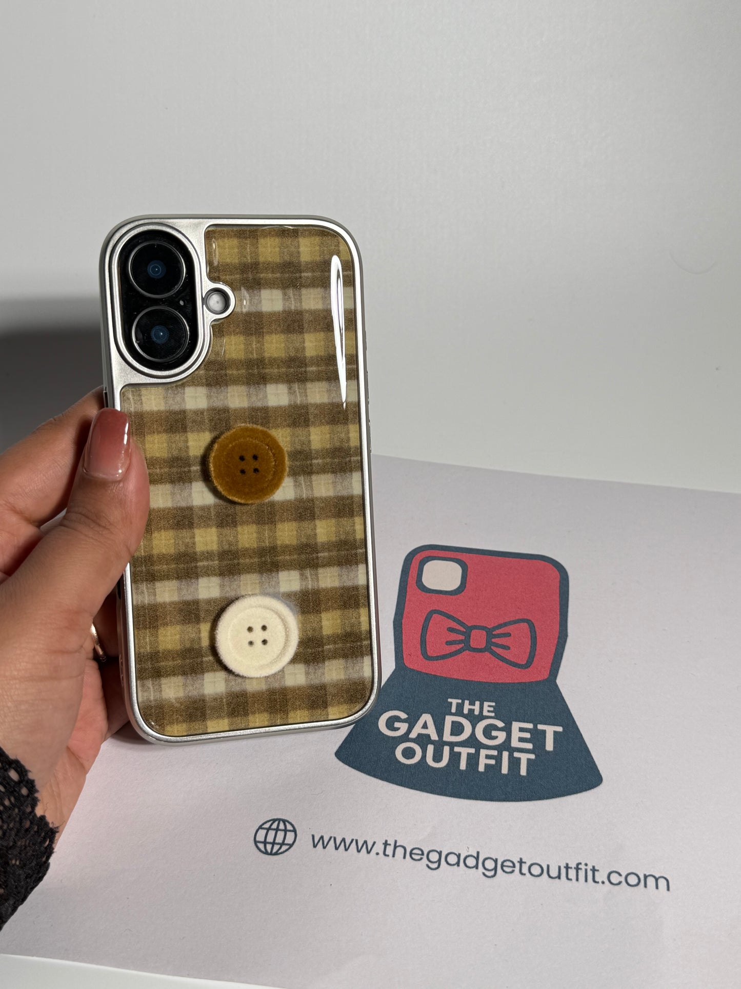 A hand holds a clear phone case featuring a tan and brown plaid pattern with two decorative buttons on the back, placed over a white surface with a 'The Gadget Outfit' logo and website printed below.